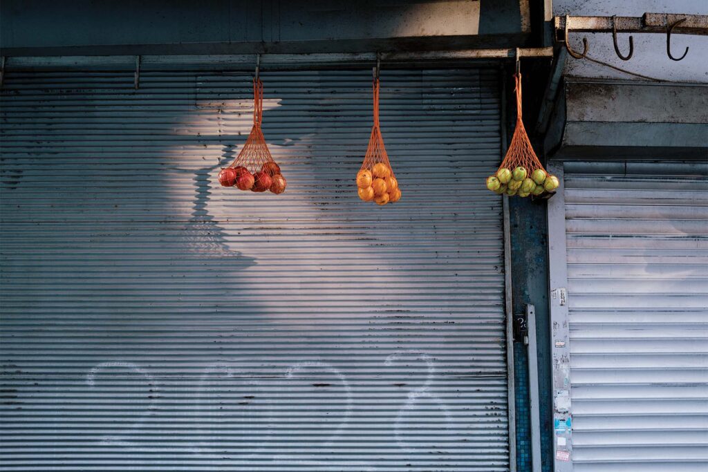 Three nets, each filled with different types of fruit, hang from the ceiling in front of a closed metal shutter. The nets contain, from left to right, red onions, oranges, and green apples. The scene is illuminated by a soft light, highlighting the textures of the fruits and the metal surface behind.