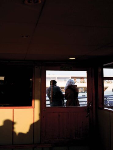 Interior of a ferry with two people at the rear window looking out at the harbor during sunset, creating silhouettes against the brightly lit background.