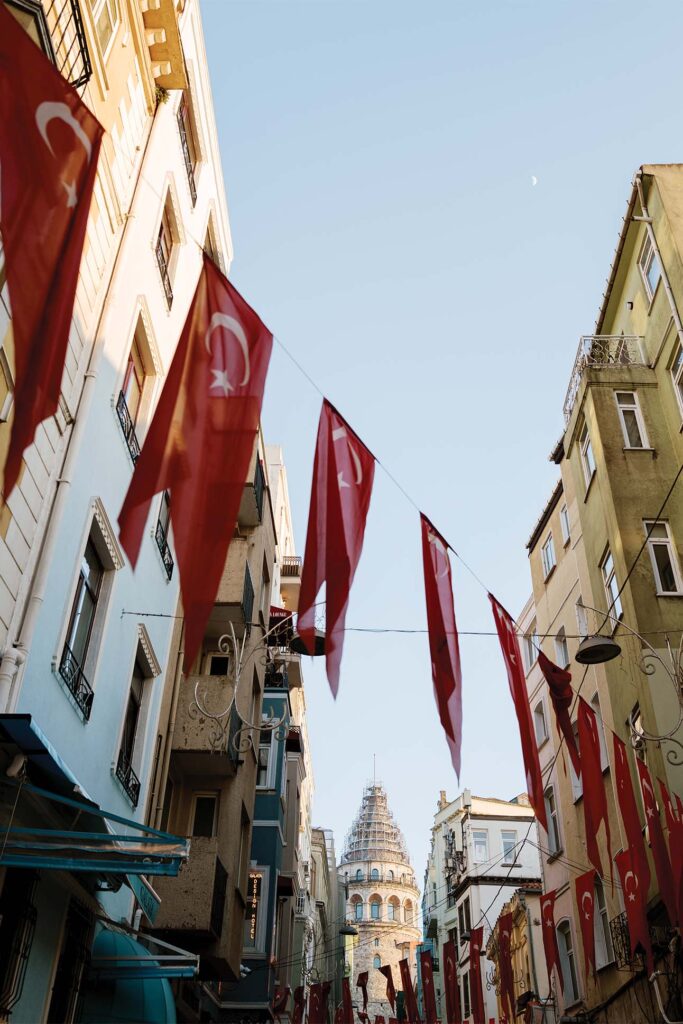 Red Turkish flags hanging between narrow buildings on a sunny day, with a historic domed building in the background and a small crescent moon visible in the sky.
