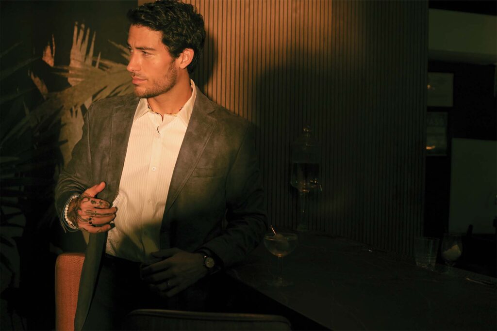 A man, in menswear fashion, wearing a gray suit jacket and white shirt sits at a bar, looking off to the side with a thoughtful expression. There's a cocktail glass on the bar in front of him. The setting is dimly lit with warm, ambient lighting.