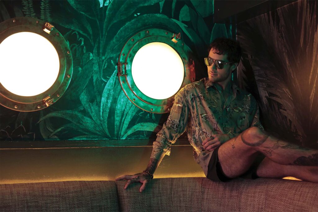 Man in menswear fashion, wearing a tropical print shirt and sunglasses sitting leisurely with a backdrop featuring two large, illuminated circular windows and vibrant green foliage patterns.