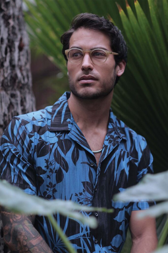 A man with glasses, in menswear fashion, wearing a floral blue shirt stands thoughtfully among tropical foliage. He has a light stubble beard and visible tattoos on his arm.