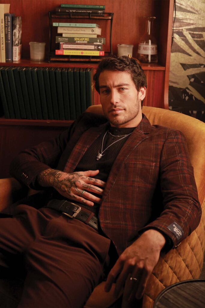 Man with curly hair and tattoos lounging in a mustard-colored armchair in a cozy room, wearing a plaid blazer over a dark shirt. He looks contemplative, with one hand resting on his lap displaying detailed tattoos. The background features a bookshelf filled with various books.