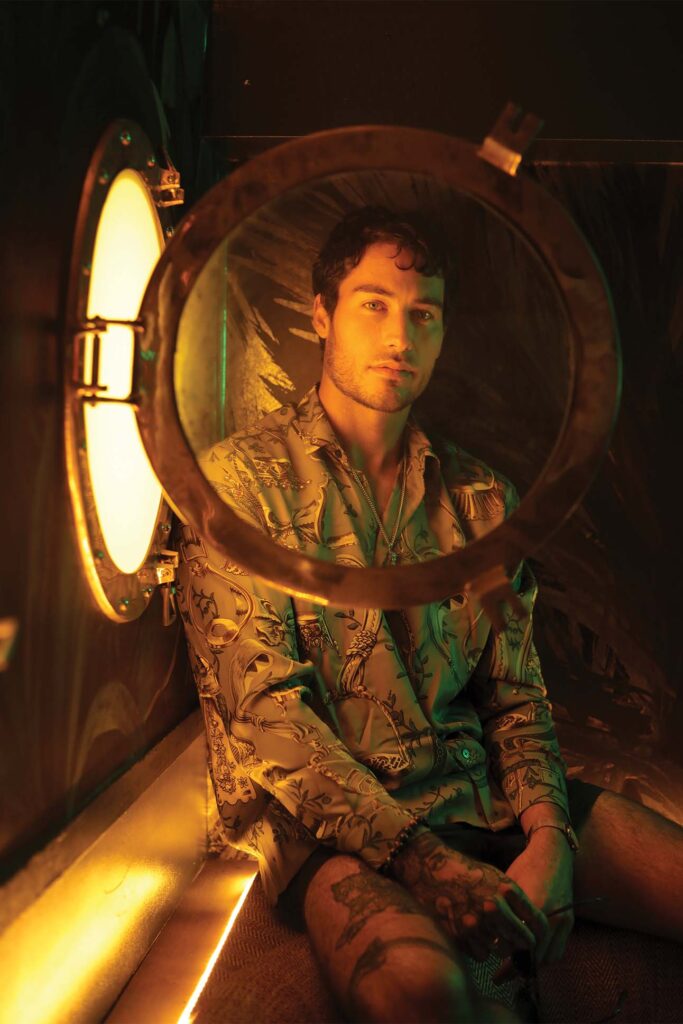 A man seated in a reflective gold room lit by a yellow-orange glow, looking intently at the camera through a circular, porthole-like opening in the foreground. He is wearing a patterned green shirt and has visible tattoos on his arms and legs.