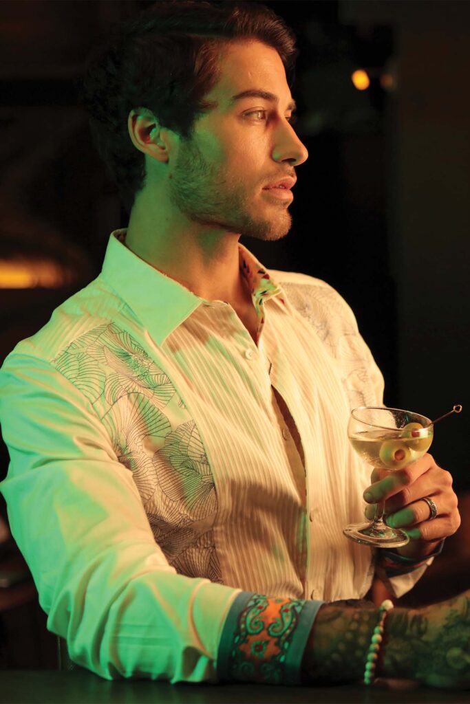 A man in a stylish bar setting, holding a martini glass with olives. He is gazing to the side with a contemplative expression. He is wearing a white shirt with a leaf pattern and green lighting highlights his features and shirt.