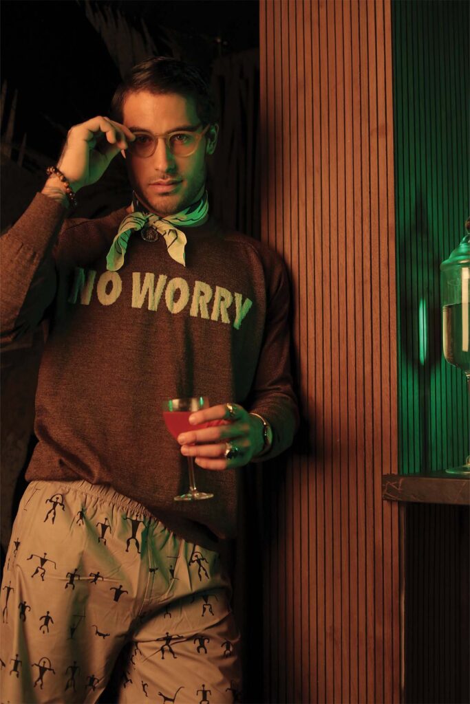 A stylish man in glasses adjusts his glasses with one hand and holds a cocktail in the other. He wears a sweater with the words "NO WORRY", a green neck scarf, and printed trousers. The setting has warm lighting and is partially lined with wooden slats.