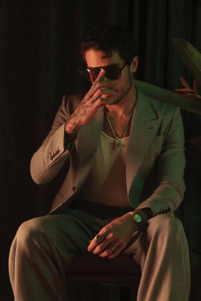 A stylish man, in menswear fashion, wearing sunglasses and a patterned suit sits on a stool in a dimly lit room, his hand partially covering his face. He has tattoos visible on his fingers, and he accessorizes with necklaces and a green watch.
