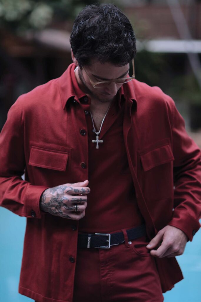 A stylish man, in menswear fashion, wearing a red shirt and jacket with a hand covered in tattoos, holding a silver cross pendant, looking downwards.
